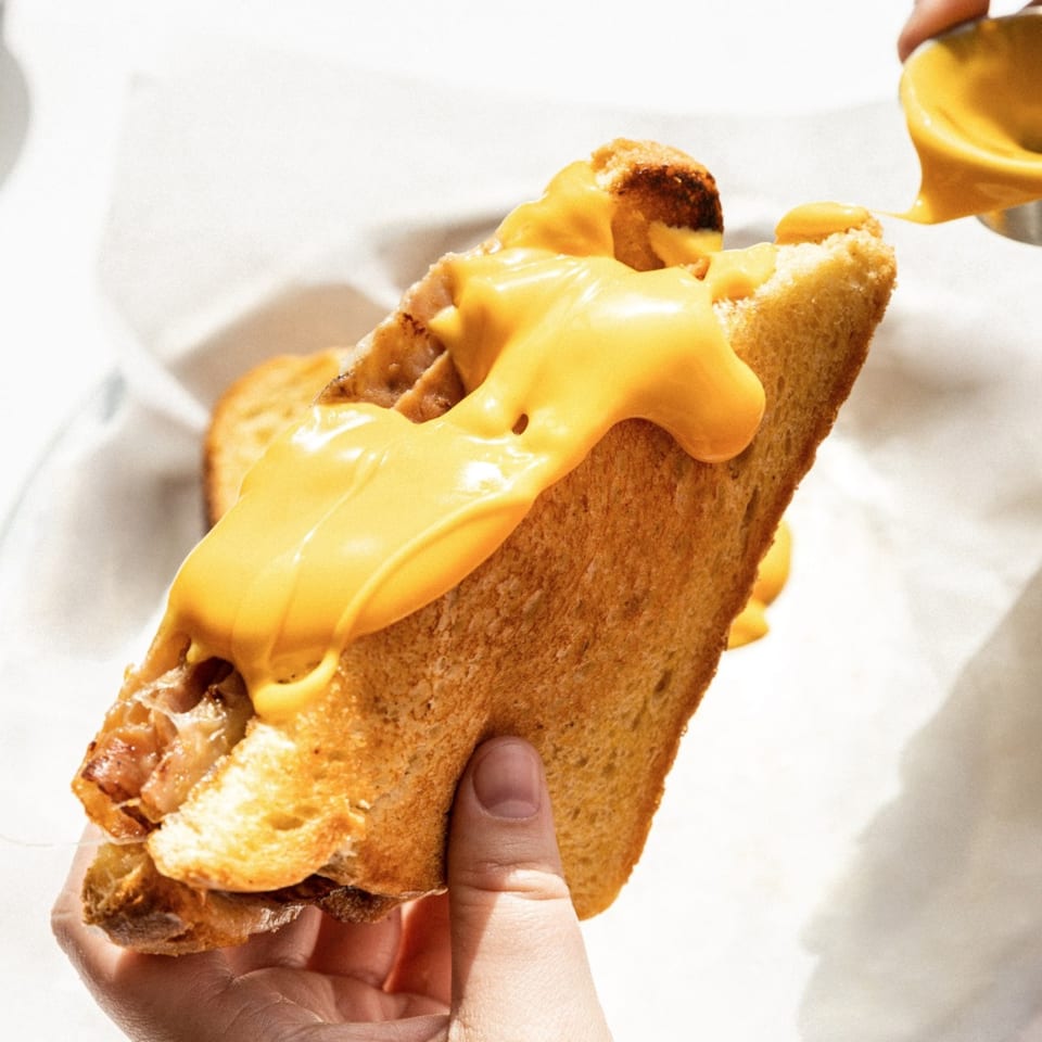Hand holding a grilled cheese sandwich with melted cheese and caramelized onions, topped with more cheese sauce.