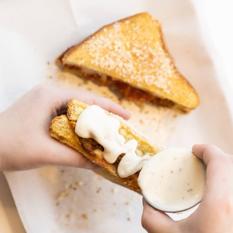 Hands squeezing white sauce onto a grilled cheese sandwich, with another sandwich in the background.
