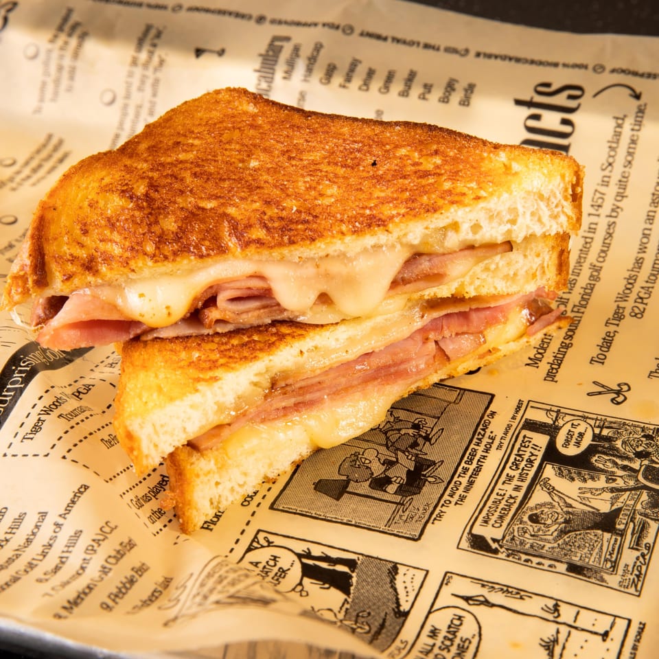 A grilled ham and cheese sandwich with melted cheese oozing out, served on newspaper-print paper.