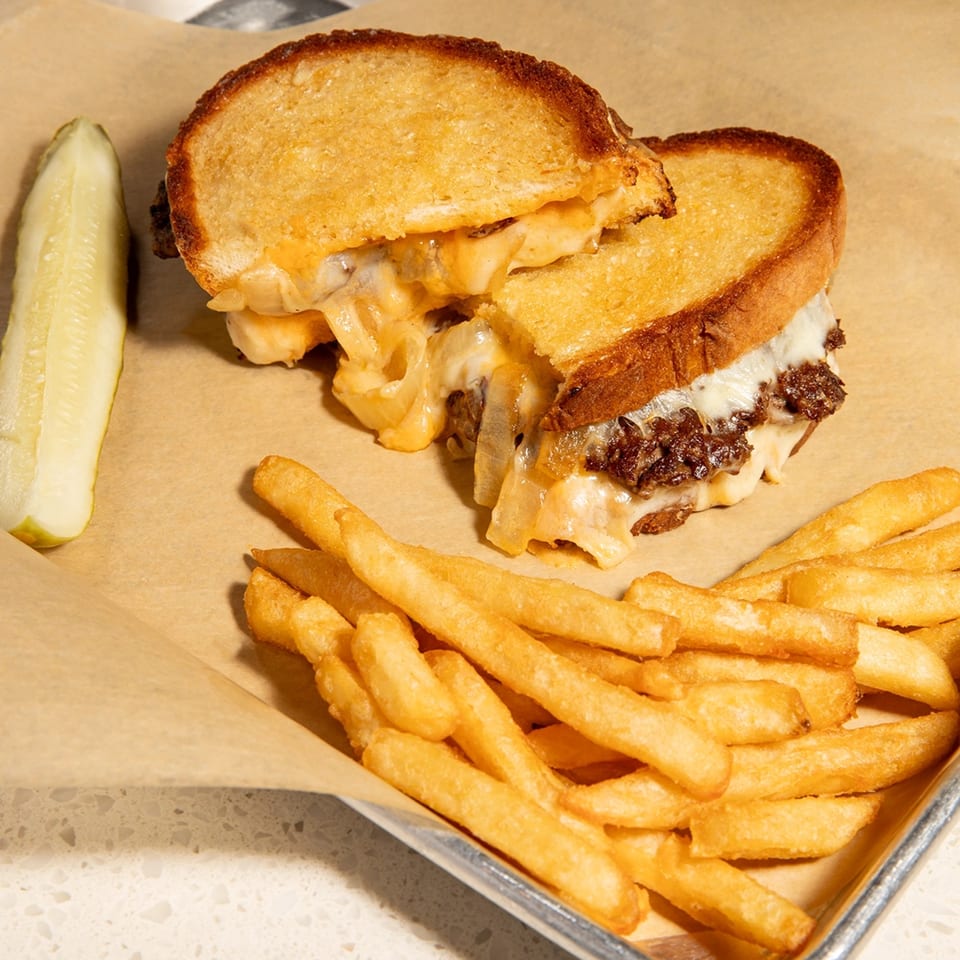 A grilled cheese sandwich with melted cheese and meat, served with fries and a pickle spear.