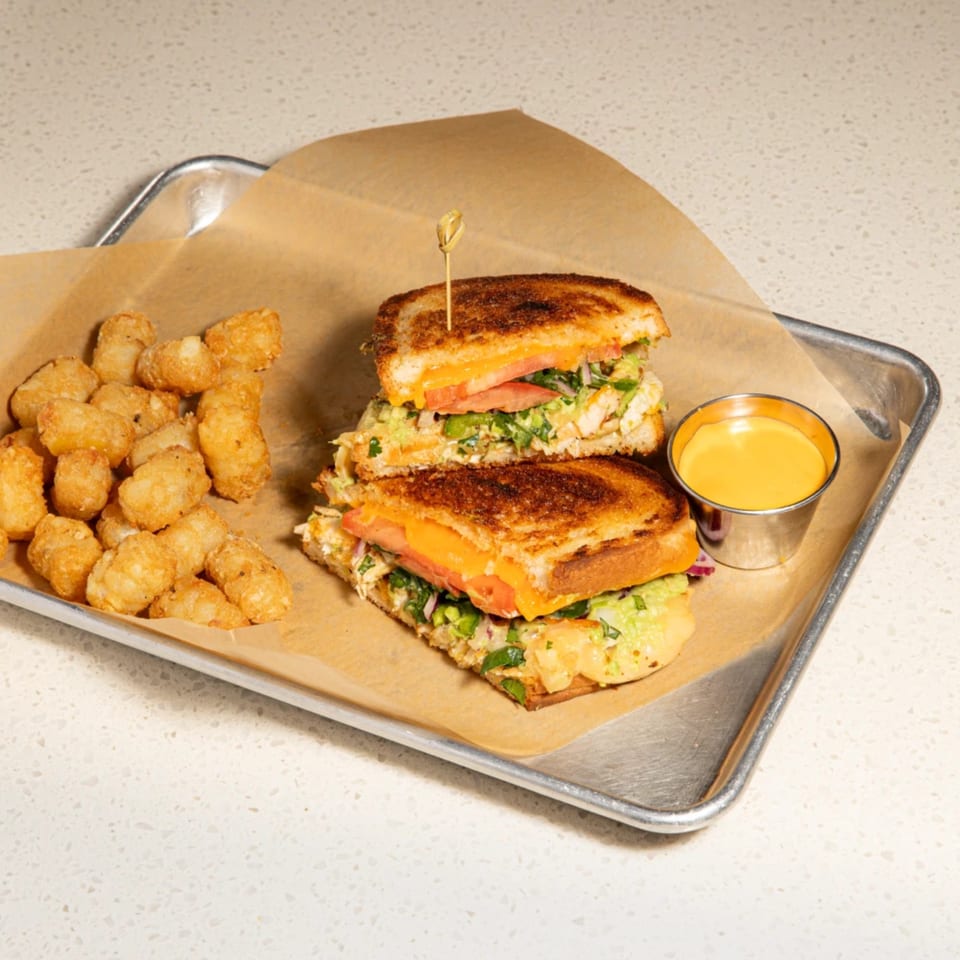 Grilled cheese sandwich halves with tomato, avocado, and chicken, tater tots, and cheese dip on a tray.