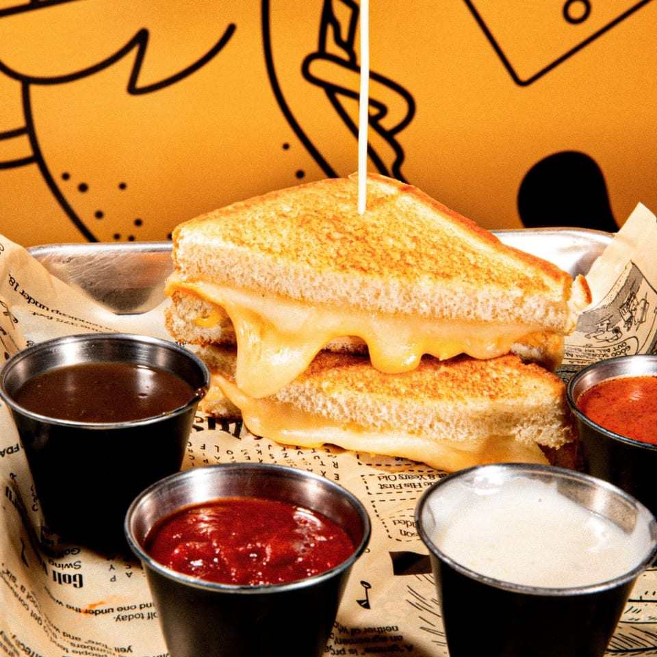 A grilled cheese sandwich with melted cheese oozing out, served with four dipping sauces on a metal tray.