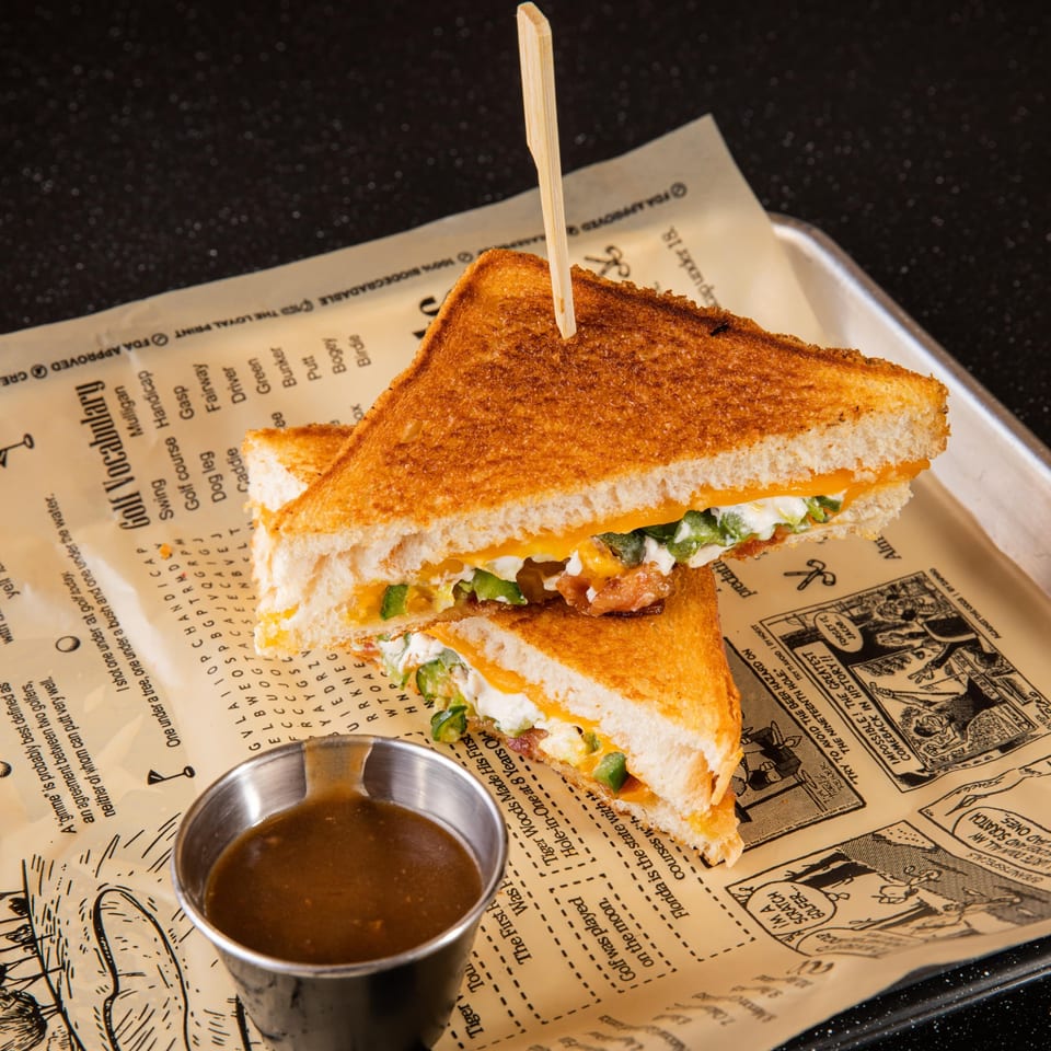 A grilled cheese sandwich with jalapeños and a side of dipping sauce on golf-themed parchment paper.