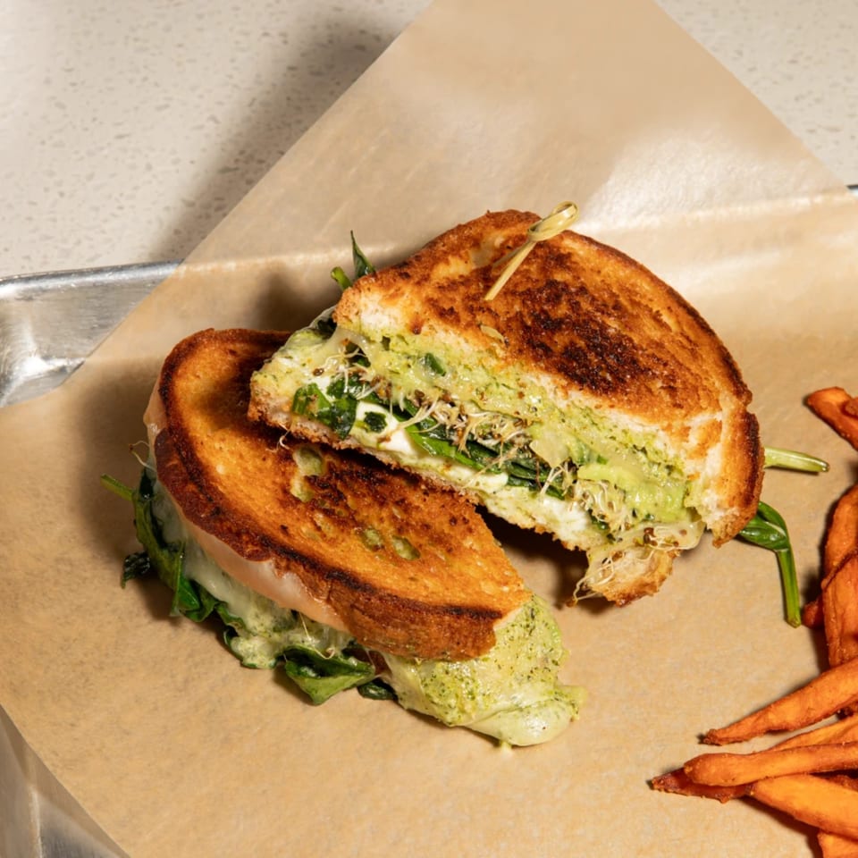 A grilled cheese sandwich, cut in half, with pesto, spinach, and sprouts, served with sweet potato fries.