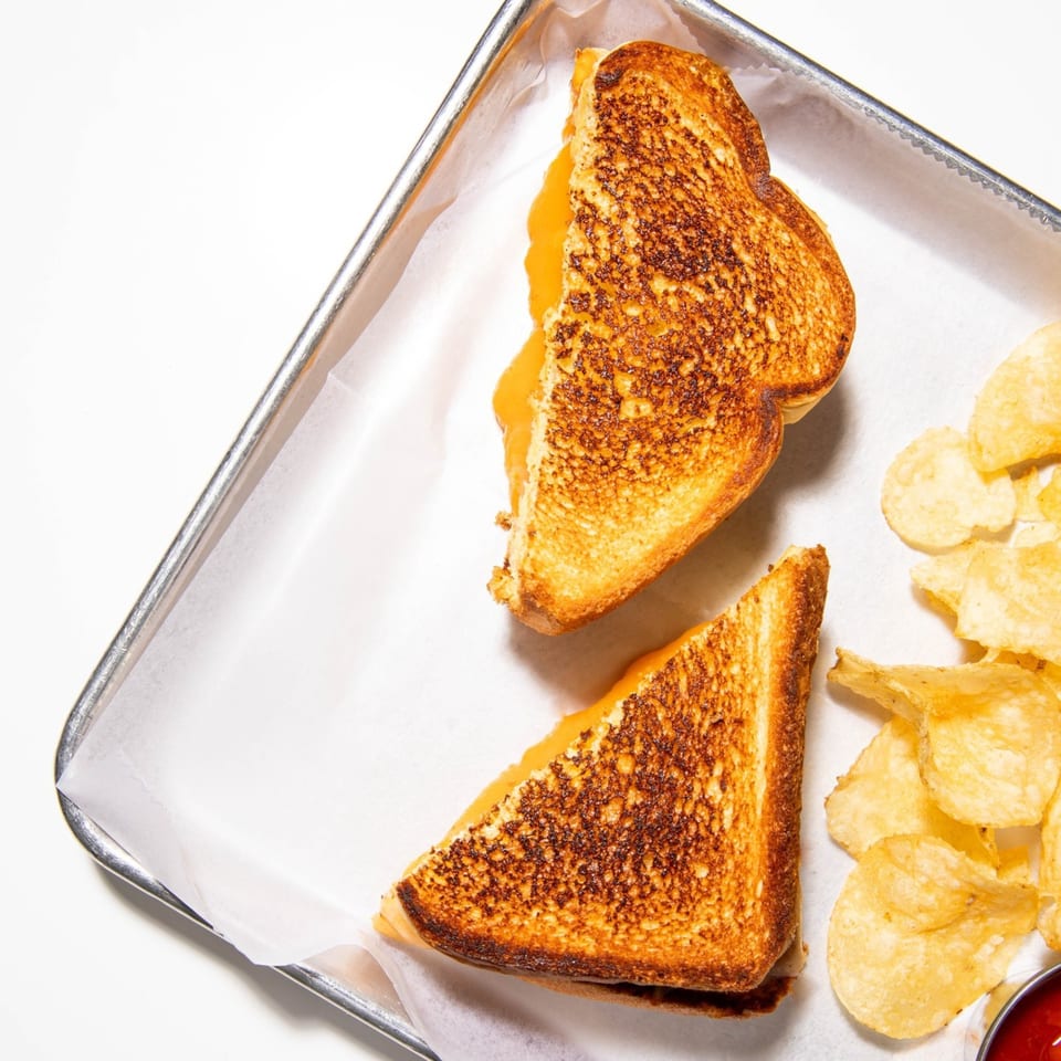 A grilled cheese sandwich, cut in half, with a side of potato chips and ketchup on a metal tray.