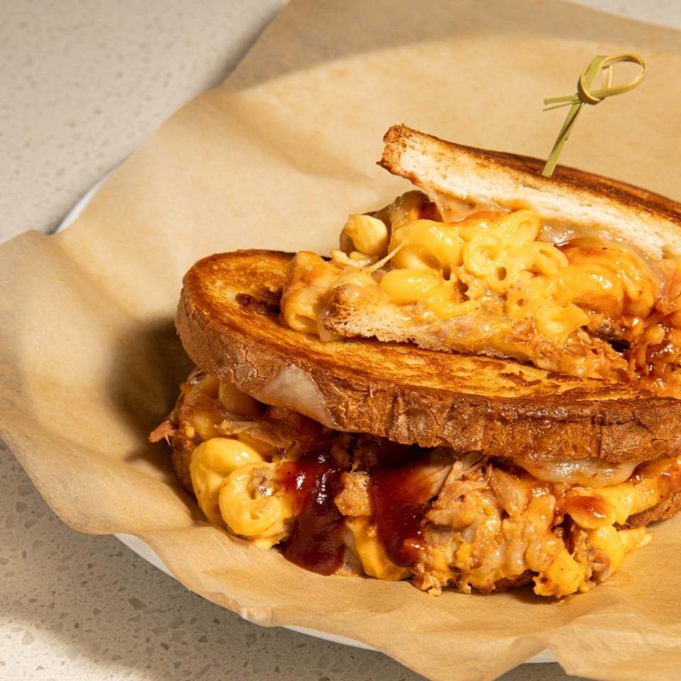 A grilled cheese sandwich with mac and cheese, pulled pork, and BBQ sauce on parchment paper.